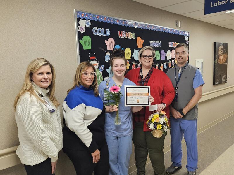 Labor and delivery nurse holds award while coworkers pose with flowers in hallway.