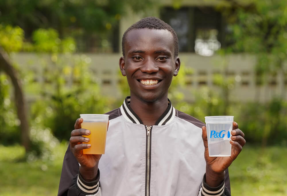 A young black man holds up two cups, one with dirty water and one with clean water in a P&G cup. He is standing outside.