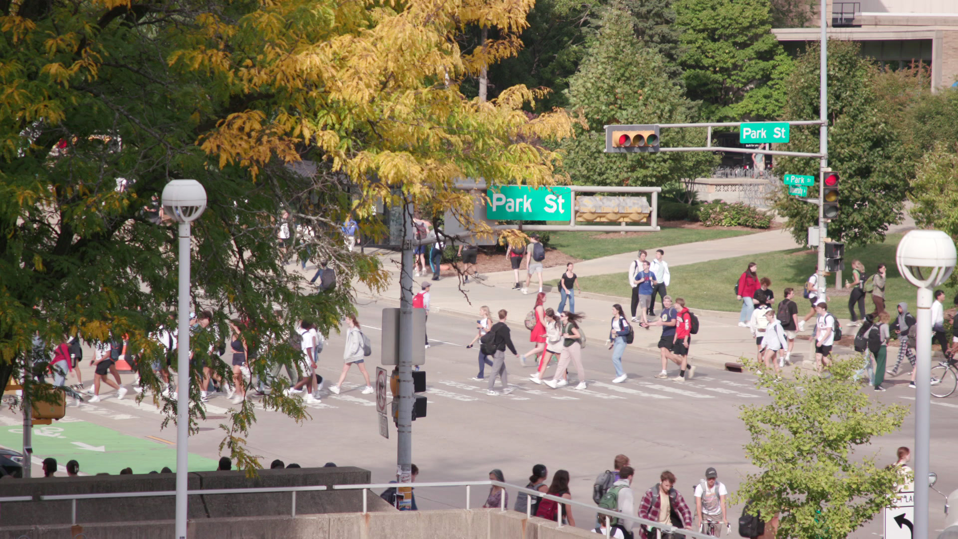 Pedestrians walk on sidewalks and across crosswalks at the intersection of two streets, with signs attached to traffic signals marking "Park St" and "University Ave," with buildings, light poles, lawns and leaf-covered trees in the foreground and background.