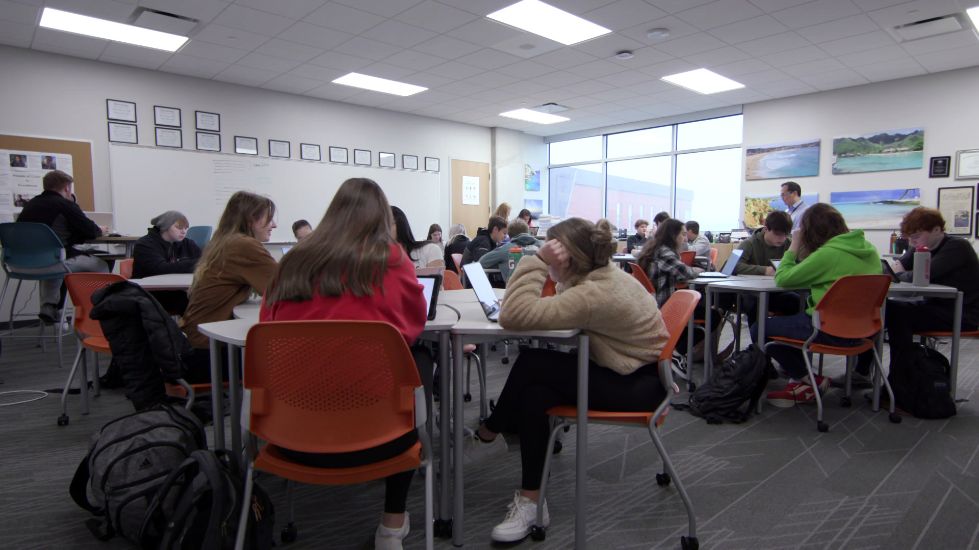 Students sit in plastic rolling chairs around tables, with multiple backpacks on a carpeted floor, with Kabby Hong standing near a back wall with photos of beach scenes and a plate glass window, in a room with fluorescent overhead light panels.