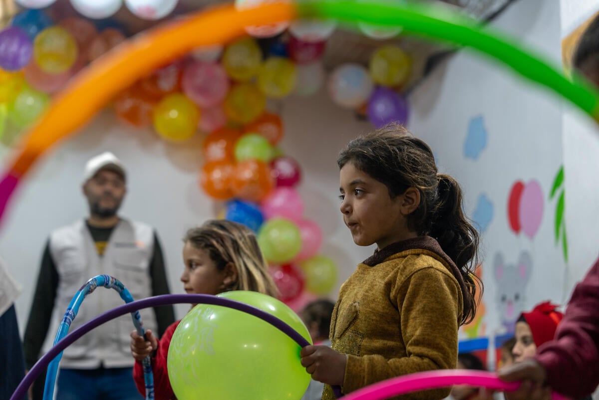 A young girl with a ponytail holds a lime-green balloon and a purple hula hoop, standing amid a cheerful scene with other children and balloons.